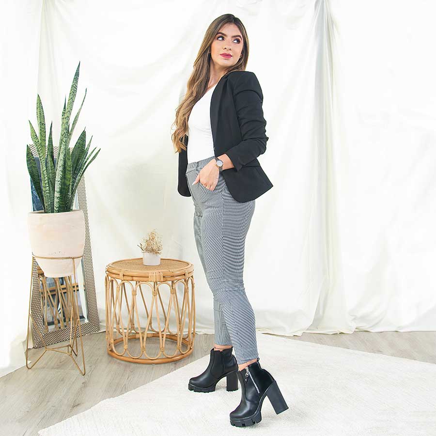 Outfit con botines negros Rebeca. Donde una mujer está luciendo unos zapatos de cuero sintético con accesorios plateados como herrajes y cierre; tiene unos jeans gris, una blusa blanca y una chaqueta negra. Además, está parada en un tapete beige y detrás de ella hay una mesa de madera con un florero blanco que tiene unas flores marchitas dentro, al lado está una maceta beige con unas plantas verdes y un espejo rectangular. Todo eso está sobre un fondo de tela beige y un tapete .