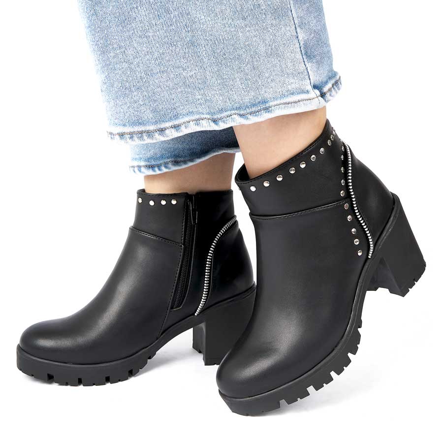 Botines para dama 262, de color negro. Están fabricados en cuero sintético, son de caña media y tacón medio. Y tienen accesorios plateados como taches, herrajes y cierre. Los zapatos los lleva calzados una modelo que lleva puestos unos jeans y está parada sobre un fondo blanco.