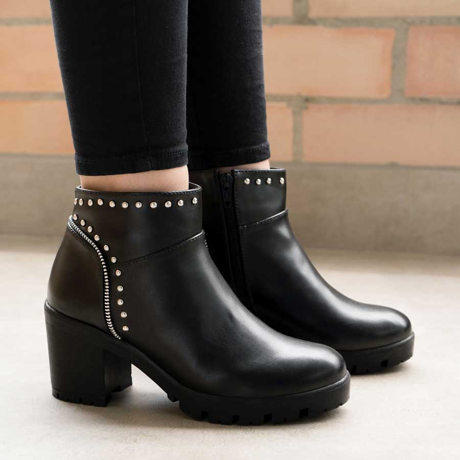 Botines negros 262 calzados por una dama. Los zapatos son en cuero sintético y tienen accesorios plateados como taches y cierres; la mujer tiene un outfit con un pantalon negro pegado. Además, está  parada en una superficie de piso claro. En la parte de atrás se ve un paredón de ladrillos.