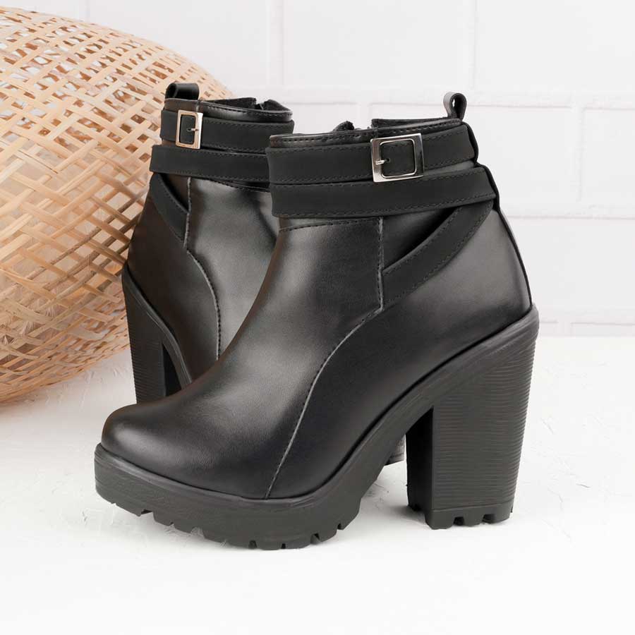 Botines para dama 286, de color negro. Están fabricados en cuero sintético, son de caña media y tacón alto. Y tienen accesorios plateados como correas y cierre. Los zapatos están de perfil ubicados uno al lado del otro y hay una maya de madera en la parte de atrás. Todo esto ubicado sobre un fondo de madera blanco. 