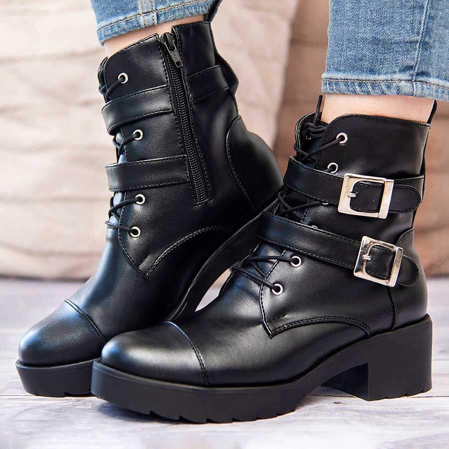 Botines negros A92 calzados por una dama. Los zapatos son en cuero sintético y tienen accesorios plateados como cordones, correas y cierre; la mujer tiene un outfit con un jean azul claro. Además, está parada en una superficie de madera gris. En la parte de atrás se ve un fondo con unos cojines color rosado. 