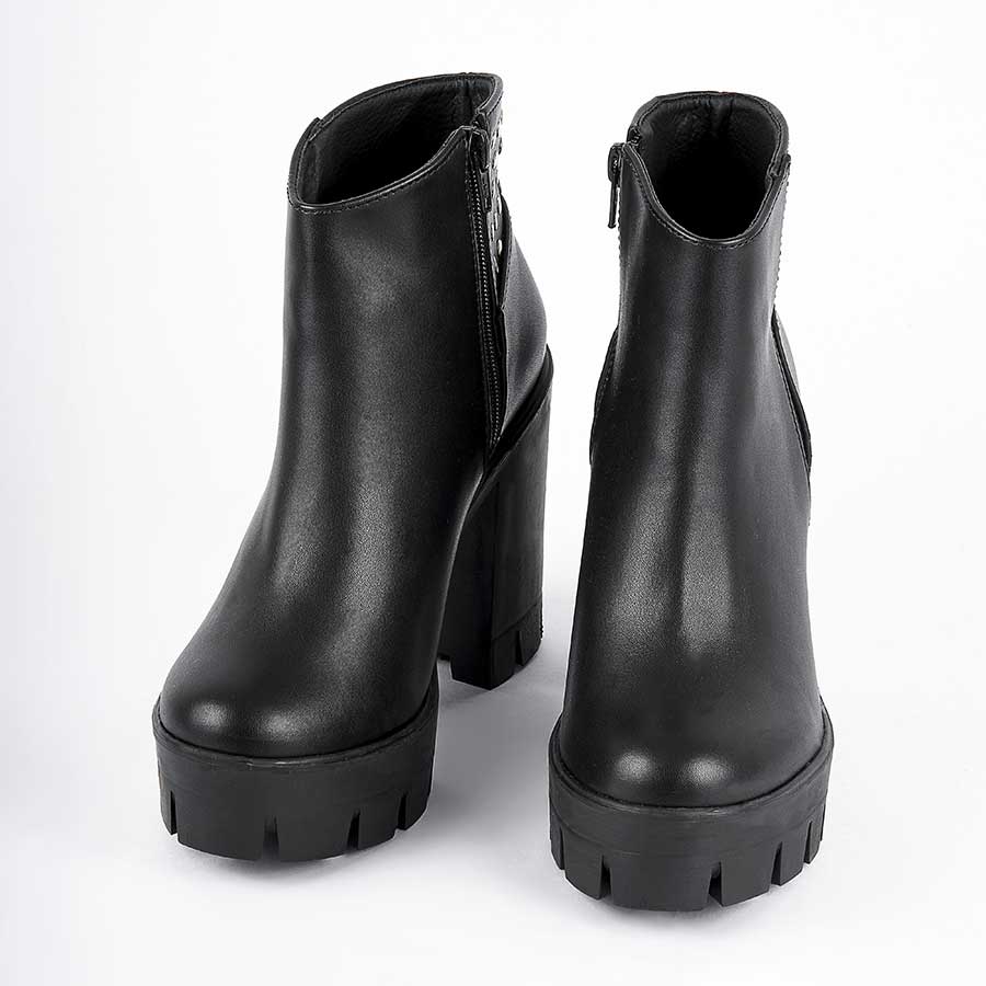 Botines para mujer dominik, de color negro. Están fabricados en cuero sintético, son de caña media y tacón alto. Y tienen accesorios plateados como taches y cierre.  Los zapatos están de frente y puestos sobre un fondo blanco.