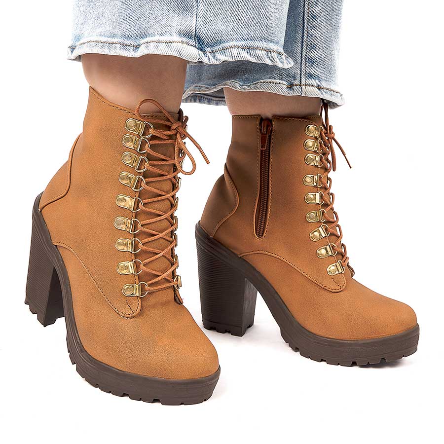 Botines para dama olivia, de color miel. Están fabricados en cuero sintético, son de caña media y tacón alto. Y tienen accesorios plateados como cordones y cierre. Los zapatos los lleva calzados una modelo que lleva puestos unos jeans y está parada sobre un fondo blanco.